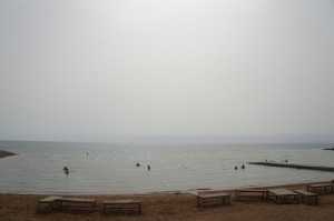 The image shows a beach scene with several empty wooden lounge chairs on the sandy shore. In the background, there is a calm body of water with a few people swimming and floating. The sky is overcast, giving the scene a muted, serene atmosphere.