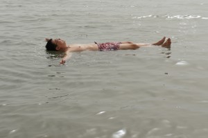 A person is floating on their back in a body of water, with arms and legs spread out. The water appears calm and the person is wearing swim trunks. The scene suggests a relaxed and peaceful moment.