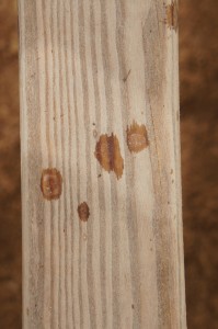 A close-up image of a wooden plank with visible grain patterns and a few dark brown knots and stains. The background is blurred and appears to be a similar wooden texture.