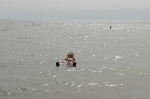 A person is floating on their back in a large body of water, possibly a sea or lake. The water appears calm, and the person is relaxed with their legs and arms spread out. The background shows a hazy horizon with distant land barely visible. The overall scene is serene and peaceful.