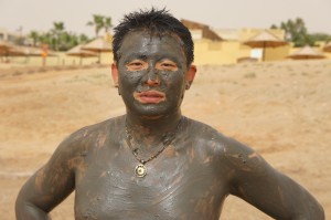 A person is standing outdoors with their face and upper body covered in mud. They are wearing a necklace and have short, dark hair. The background shows a dry, sandy landscape with some buildings and trees in the distance.