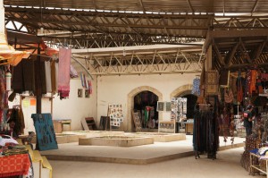 The image shows an indoor market with various stalls displaying colorful textiles, clothing, and other goods. The market has a high ceiling with metal beams and a central open space. The stalls are filled with hanging fabrics, bags, and other items for sale. The background features arched doorways and walls with some decorations. The overall atmosphere is vibrant and busy, typical of a traditional market setting.