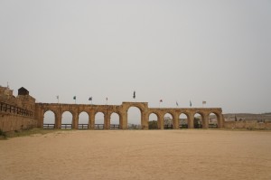 The image shows an ancient stone structure with multiple arches, likely part of a historical site or fortification. The structure is set against a clear sky and has a sandy ground in the foreground. Several flags are visible on top of the structure, and there is a statue or monument in the center of the arches.