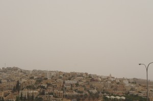 The image shows a hilly landscape densely populated with buildings. The sky is overcast, giving the scene a muted, hazy appearance. There is a streetlight visible on the right side of the image. The buildings are mostly light-colored and appear to be closely packed together.