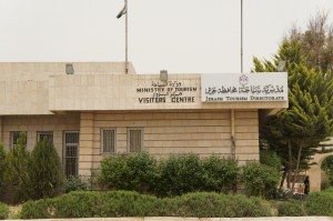 The image shows a building with a sign that reads "Ministry of Tourism Visitors Centre" in both English and Arabic. The building is surrounded by greenery, including bushes and trees. There are two flagpoles on the roof, one of which has a flag. The structure appears to be made of light-colored stone or concrete.