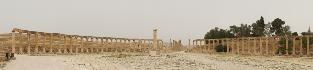 The image shows the ruins of an ancient Roman colonnade, with a series of tall stone columns arranged in a semi-circular formation. The ground is paved with large, uneven stones, and there is a single column standing in the center of the open area. The background features a few trees and a hazy sky. The overall scene appears to be an archaeological site, likely an ancient city or forum.