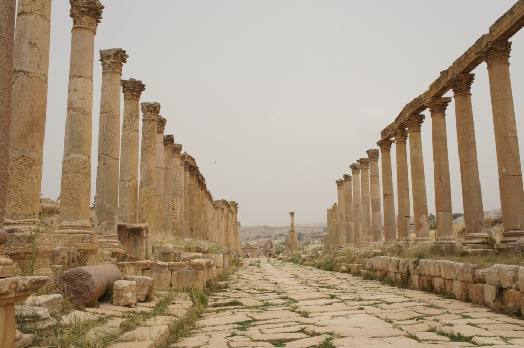 Alt text: A view of an ancient Roman colonnaded street with tall stone columns on both sides. The columns are weathered and some are partially broken. The street is paved with large stone slabs, and there is grass growing between the stones. The sky is overcast, and the background shows a distant landscape with more ruins and hills.