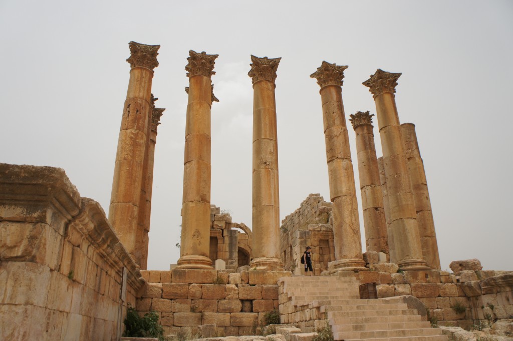 The image shows the ruins of an ancient structure with several tall, weathered columns. The columns are made of stone and have ornate capitals at the top. They are arranged in a row, and some of them are partially broken. In the background, there are remnants of additional stone structures. In the foreground, there are stone steps leading up to the columns, and a person is visible standing near the top of the steps. The sky is overcast, giving the scene a muted, historical atmosphere.