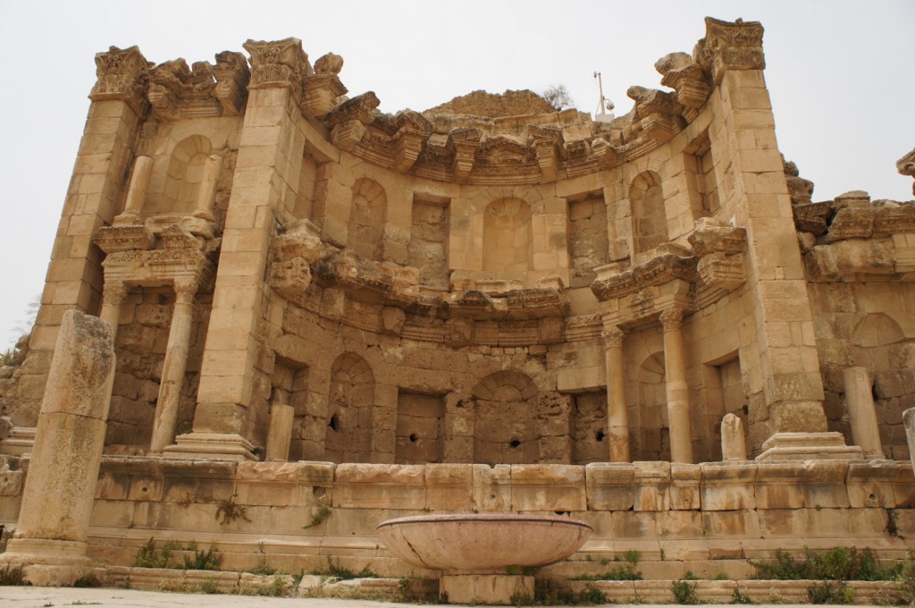 Alt text: A large, ancient stone structure with multiple levels of arches and columns. The facade is weathered and partially damaged, with intricate carvings and architectural details. In front of the structure, there is a large, empty stone basin. The ground around the structure is uneven and has patches of grass and small plants. The sky above is overcast.