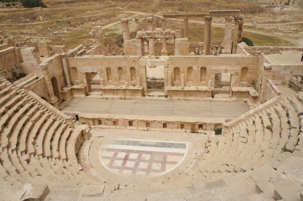 The image shows an ancient Roman amphitheater with a semi-circular arrangement of stone seating surrounding a central stage area. The stage is backed by a partially intact stone wall with columns and arches, indicating the remains of a grand architectural structure. The surrounding landscape is arid and rocky, with sparse vegetation. The amphitheater appears to be in a state of ruin, but still retains much of its original structure.