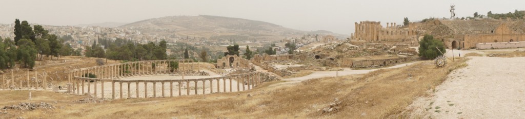 The image shows a panoramic view of ancient ruins in a dry, hilly landscape. The ruins include a large, semi-circular colonnade with numerous columns, and various other stone structures and remnants of buildings scattered across the area. In the background, there are more hills and a distant view of a modern town or city. The sky is overcast, giving the scene a muted, beige tone. Sparse vegetation is visible, with a few trees and patches of grass.