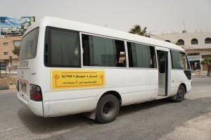 A white minibus is parked on the side of a road. The bus has a yellow sign with Arabic text on its side. The bus door is open, and there are buildings and palm trees in the background. The sky appears to be overcast.