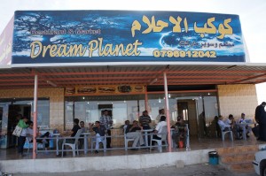 The image shows the exterior of a restaurant named "Dream Planet." The restaurant has a large sign with both Arabic and English text, and a phone number is displayed. There are several people sitting at tables outside the restaurant, some of whom appear to be eating or drinking. The building has a covered patio area with a few tables and chairs. The overall atmosphere seems casual and relaxed.