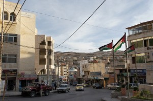 A street scene in a small town with several buildings on either side. There are cars parked along the street and a few driving down the road. Three flags are prominently displayed on poles in the middle of the image. The sky is partly cloudy, and there are hills in the background. The buildings have a mix of modern and traditional architectural styles. Power lines are visible overhead.