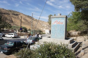 The image shows a stone monument with the word "Petra" written on it in red letters. The monument is situated in an outdoor area with a few cars parked nearby. There are some people standing around the cars. The background features a hilly landscape with sparse vegetation and a clear blue sky. Some trees and bushes are also visible in the foreground.