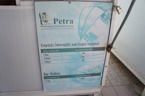 The image shows a signboard for Petra, detailing the entry ticket prices for tourists (overnight and cruise visitors) and day visitors. The sign includes the following information:

- For tourists (overnight and cruise visitors):
  - 1 Day: 50 JD
  - 2 Days: 55 JD
  - 3 Days: 60 JD

- For day visitors:
  - Entry Ticket: 90 JD

The sign is mounted on a wall, and the background features a light blue design with images of tickets.