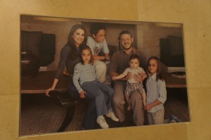 A family portrait featuring two adults and four children. The adults are seated in the center, with the man holding a young child on his lap. The woman is sitting to the left, and three other children are positioned around them. The background appears to be a cozy indoor setting with a wooden table and cushioned seating.