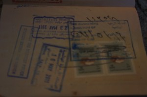 The image shows a page from a passport with multiple stamps and two visa stickers. The stamps are in various shapes and sizes, mostly rectangular, and are in blue ink. The visa stickers are placed at the bottom of the page and have some text and images on them. The page appears to be well-used, indicating multiple entries and exits from different countries.