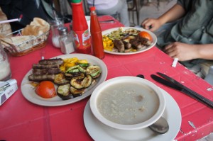 The image shows a table with a red tablecloth, on which there are two plates of food and a bowl of soup. One plate contains grilled vegetables, including zucchini and bell peppers, along with sausages and a tomato. The other plate has a similar assortment of grilled vegetables and meats. There are also condiments, including bottles of ketchup and hot sauce, and a basket of bread on the table. Two people are seated at the table, with their hands visible.