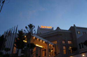 The image shows the exterior of a building with the "Marriott" sign illuminated at the top. The building appears to be a hotel, with a well-lit entrance and several flags displayed on poles to the left. The sky is clear and it seems to be either early morning or evening, as the lighting suggests a low-light condition.
