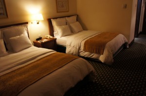 The image shows a hotel room with two neatly made double beds. Each bed has white linens and a golden-brown blanket folded at the foot. There is a nightstand between the beds with a lamp, a telephone, and some other small items. The room has a patterned carpet and framed artwork on the walls. The lighting is warm and gives the room a cozy atmosphere.