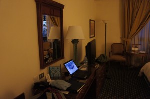 The image shows a cozy hotel room with a warm, dim lighting. There is a wooden desk with a laptop open on it, displaying a webpage. The desk also has various items scattered on it, including clothes and a water bottle. Above the desk is a large mirror with a wooden frame. Next to the desk is a flat-screen TV on a stand. In the background, there is a tall floor lamp, a chair with a cushion, and a small table with a few items on it. The room has a carpeted floor and curtains covering the window.