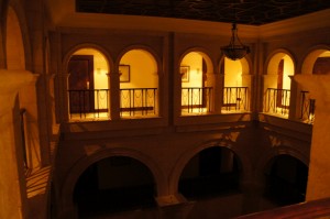 The image shows an interior view of a building with an architectural design featuring multiple arches. The scene is dimly lit with warm, yellow lighting. There are two levels visible, each with a series of arches and railings. The upper level has doors and framed pictures on the walls, while the lower level is darker and less detailed. A chandelier hangs from the ceiling in the center of the image.