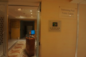 The image shows the entrance to a facility with signs indicating the presence of a swimming pool and public toilets. There is a reception desk with a computer monitor on it. The wall has a sign that reads "LOOFAH Turkish Bath." The area is well-lit with ceiling lights, and the floor has a patterned design.