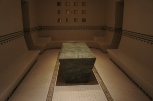 The image shows the interior of a steam room or sauna. The room has tiled walls and floors with a central stone or marble table. There are built-in benches along the walls on either side of the table. The lighting is dim, creating a relaxing and serene atmosphere. The walls have a decorative pattern with small square tiles near the ceiling.