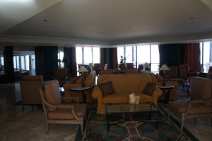 The image shows a spacious, well-lit lounge area with multiple seating arrangements. There are several armchairs and sofas, including a central yellow sofa with a glass coffee table in front of it. The room has large windows that allow natural light to flood in, and there are various side tables with lamps and decorative items. The floor is covered with a patterned rug, and the overall decor is elegant and comfortable.