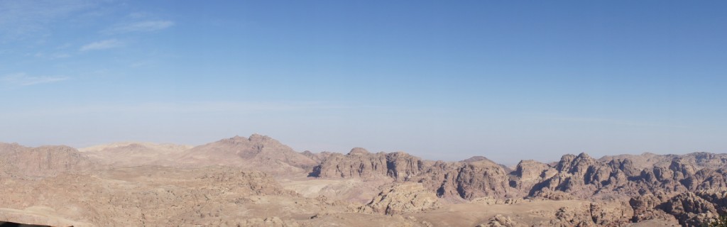 A panoramic view of a rocky desert landscape with rugged mountains under a clear blue sky. The terrain is arid and barren, with various shades of brown and tan. The horizon is visible in the distance, and the sky is mostly clear with a few wispy clouds.