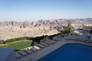 The image shows a scenic view of a swimming pool area with several lounge chairs arranged around it. Beyond the pool, there is a lush green lawn bordered by bushes. In the background, there is a vast, rocky desert landscape with rugged mountains under a clear blue sky. The setting appears to be a serene and luxurious outdoor space, likely part of a resort or hotel.