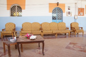 The image shows a room with a set of yellow upholstered chairs and a sofa arranged against a wall. The wall is painted in two colors: the lower half is light blue, and the upper half is white with orange accents around the windows. There are two arched windows with metal grilles. In front of the seating area, there is a wooden coffee table with a few items on it, including a small bouquet of flowers and a white object. The floor has a patterned tile design. The room appears to be well-lit and has a simple, cozy atmosphere.