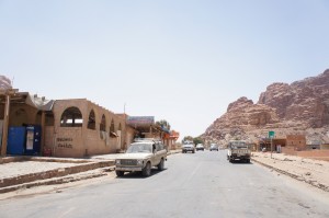 Alt text: A street scene in a desert town with a few vehicles, including a white pickup truck, parked along the road. On the left side, there is a building with arched windows and a sign that reads "Wadi Rum." In the background, there are rocky desert mountains under a clear blue sky.