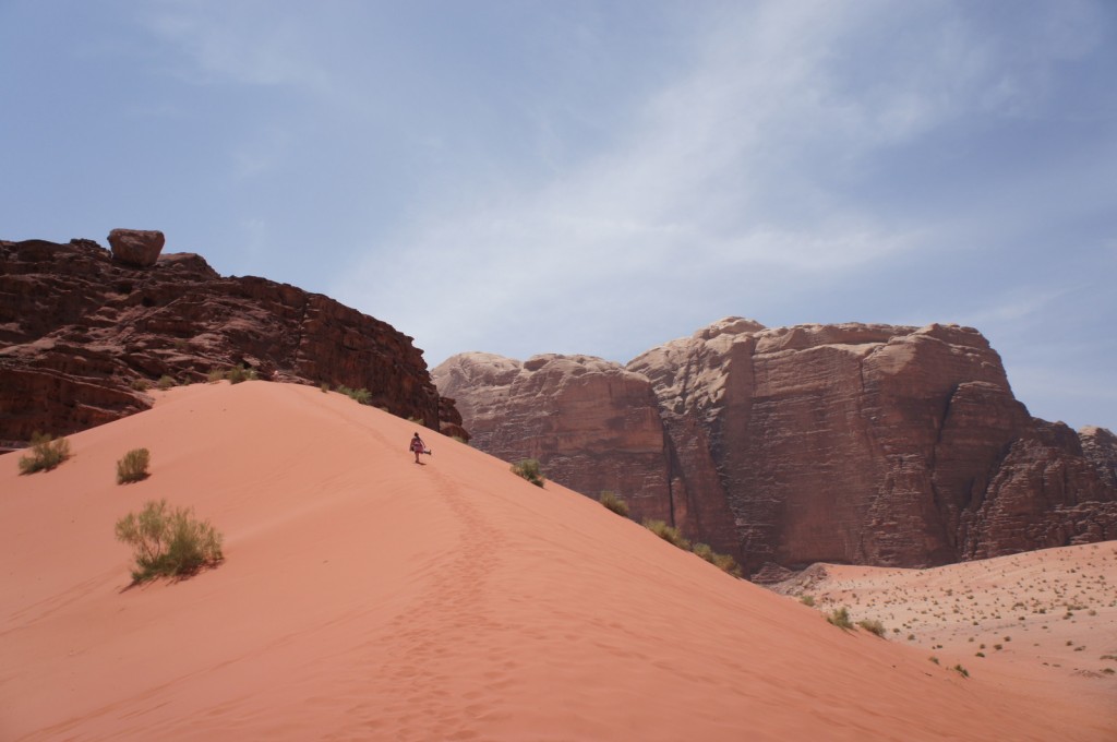 A person is walking up a large sand dune in a desert landscape. The sand is a reddish color, and there are some small bushes scattered on the dune. In the background, there are tall, rugged rock formations and a clear blue sky.