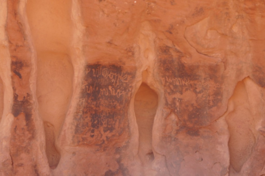 The image shows a close-up view of a rock face with ancient petroglyphs or carvings. The rock is reddish-brown in color, and the carvings appear to be symbols or writing etched into the surface. The carvings are darker in color, contrasting with the lighter rock. The rock surface has a rough texture with natural grooves and indentations.