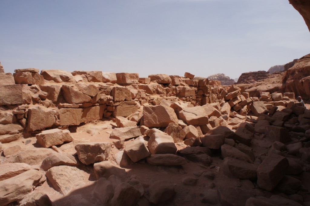 The image shows a landscape of ancient ruins made of large, weathered stone blocks. The stones are scattered and piled, suggesting the remnants of a collapsed structure. The surrounding area is arid and rocky, with a clear sky overhead. In the background, there are rocky hills or mountains. The scene appears to be in a desert or semi-desert environment.