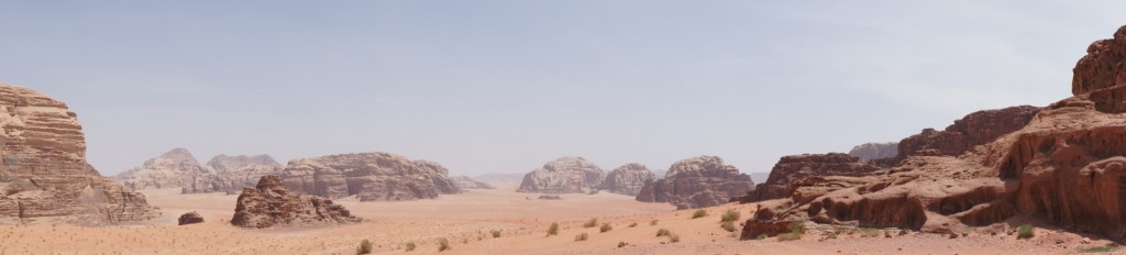 A panoramic view of a desert landscape with rocky formations and mountains in the background. The sky is clear and blue, and the ground is covered with sand and sparse vegetation. The rocky formations vary in size and shape, creating a rugged and dramatic scene.