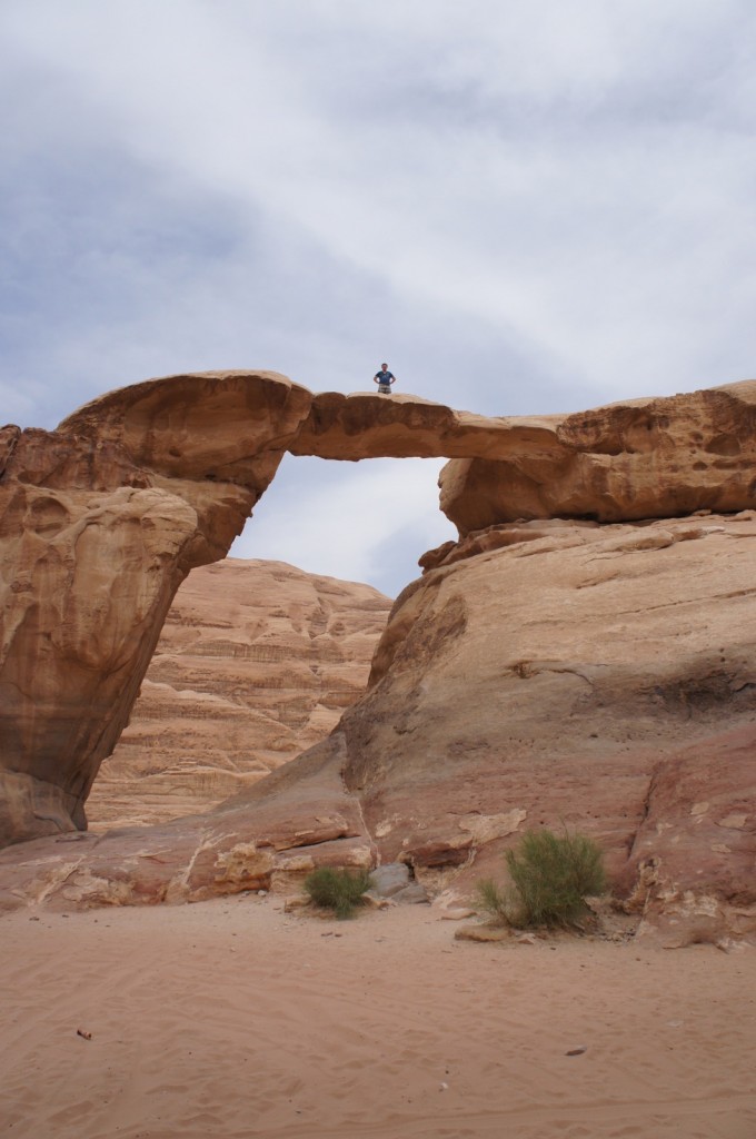 A person is standing on top of a natural rock arch in a desert landscape. The sky is partly cloudy, and the surrounding area consists of sandy terrain and rocky formations. Some small bushes are visible at the base of the rock arch.