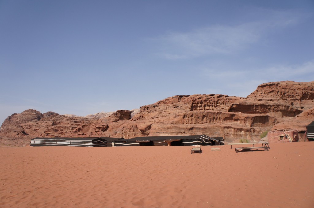 Alt text: A desert landscape with reddish sand and rocky cliffs in the background. In the foreground, there are several black and white striped tents and a few wooden benches. The sky is clear and blue.