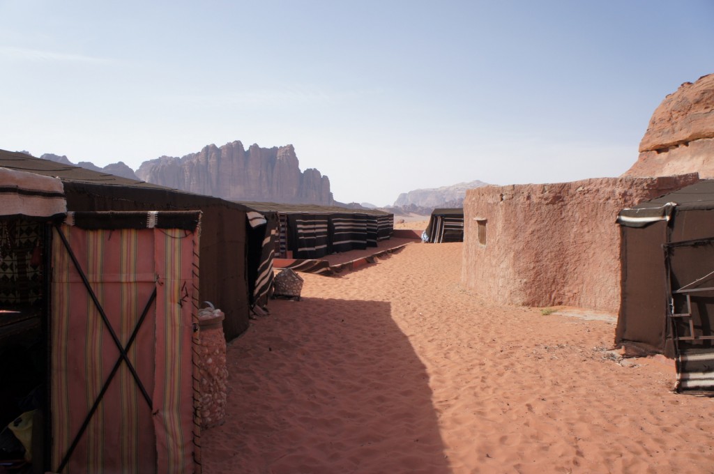 The image shows a desert scene with traditional Bedouin tents and a round stone structure. The ground is covered with reddish sand, and there are rocky mountains in the background under a clear blue sky. The tents are made of dark fabric with some striped patterns, and the area appears to be a campsite or a small settlement.