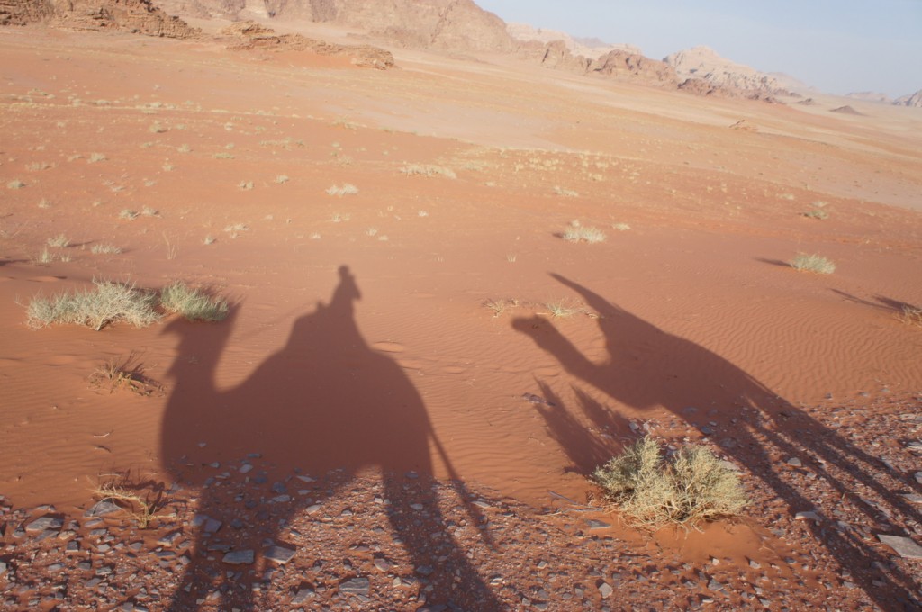 The image shows the shadows of two camels and their riders cast on the sandy desert ground. The desert landscape is vast and arid, with sparse patches of vegetation and distant rocky hills under a clear sky.