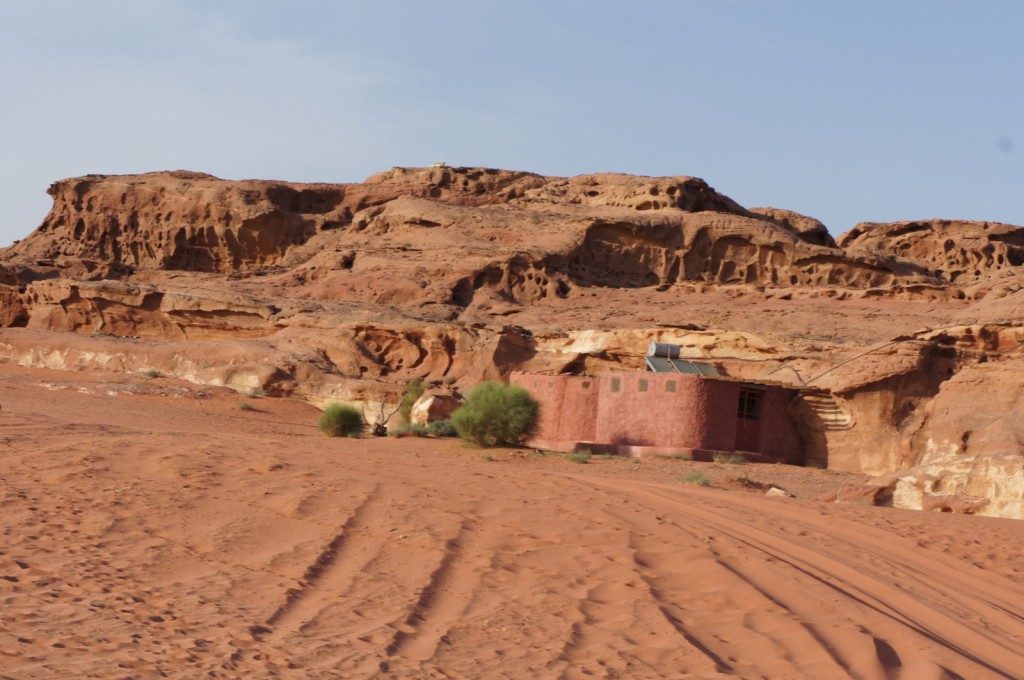 A small, pinkish building with a flat roof is nestled against a rocky, desert hillside. The surrounding landscape is arid, with reddish sand and sparse vegetation, including a few small bushes. The rocky hillside has a rugged, eroded appearance with various crevices and formations. The sky above is clear and blue.