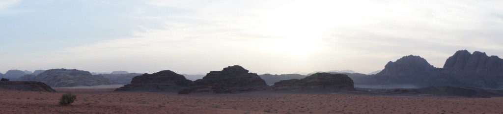 A panoramic view of a desert landscape with rocky formations and mountains in the background. The sky is partly cloudy with the sun partially obscured, casting a soft light over the scene. The ground is covered with reddish sand and sparse vegetation.