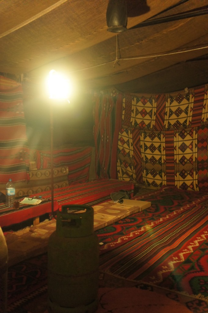 The image shows the interior of a traditional tent, possibly a Bedouin or nomadic tent. The walls and floor are covered with colorful, patterned textiles. There is a bright light source hanging from the ceiling, illuminating the space. On the floor, there are low seating arrangements with cushions and a small table. A green gas cylinder is visible in the foreground, and there are various items such as a water bottle and some papers on the table. The overall ambiance is warm and cozy.