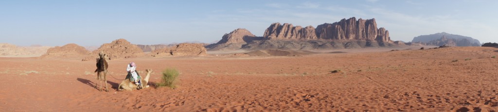A vast desert landscape with reddish sand and scattered vegetation. In the foreground, a person is sitting on the ground next to a camel, while another camel stands nearby. In the background, there are large, rugged rock formations and mountains under a clear blue sky.