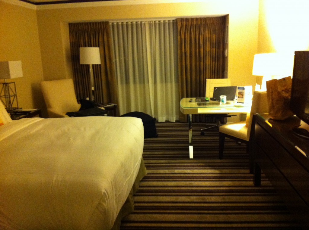 The image shows a well-lit hotel room with a modern and cozy design. There is a large bed with white linens on the left side of the room. Next to the bed, there is a nightstand with a lamp. In the background, there are floor-to-ceiling windows covered with curtains. In front of the windows, there is a desk with a chair, a laptop, and some other items on it. To the right of the desk, there is another chair and a lamp on a table. The room has a striped carpet and beige walls, creating a warm and inviting atmosphere.