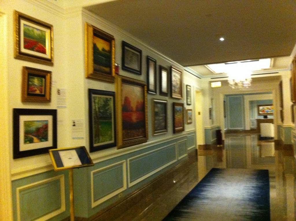 The image shows a hallway in what appears to be an art gallery or a hotel. The walls are adorned with numerous framed paintings of various sizes and styles. The hallway has a polished floor and is well-lit with ceiling lights and a chandelier. There is a stand with a sign or information board on the left side of the hallway. The overall decor is elegant, with light-colored walls and blue paneling.