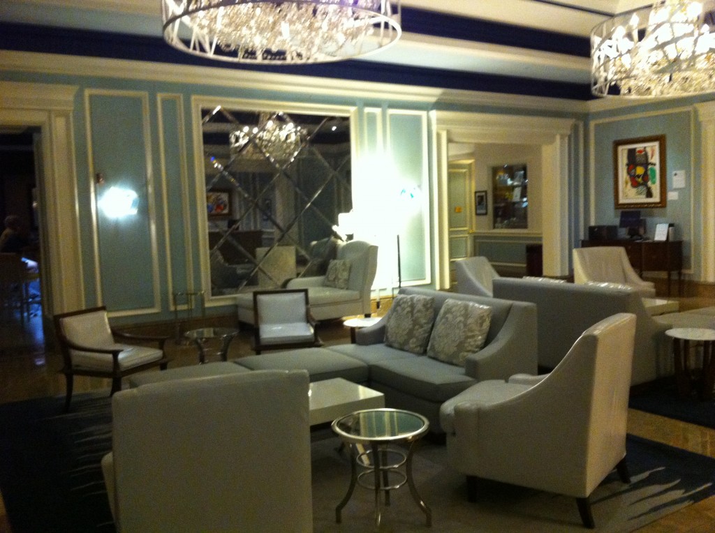 The image shows a luxurious and elegantly decorated lounge area. The room features light blue walls with white trim and is furnished with modern, light-colored chairs and sofas. There are several glass-topped tables scattered around the seating area. The ceiling has large, ornate chandeliers that provide a warm and inviting light. A large mirror with a diamond pattern is mounted on one wall, reflecting the room's decor. There is also a piece of colorful abstract art hanging on another wall. The overall ambiance of the room is sophisticated and comfortable.