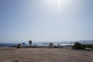 The image shows a coastal scene with a clear blue sky and bright sunlight. In the foreground, there is a sandy or dirt area leading to a small harbor or marina. Several boats and ships are visible in the water, and there are a few palm trees and some vegetation near the shore. The background features a calm sea with more ships in the distance and a faint outline of mountains or hills on the horizon.